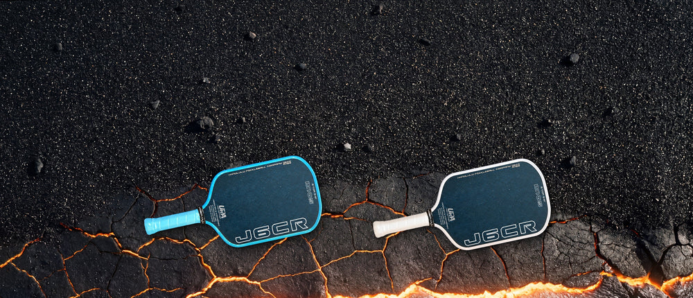Two Honolulu J6CR M105 paddles, one Blue and one White, lying on black volcanic sand where it meets glowing lava crust