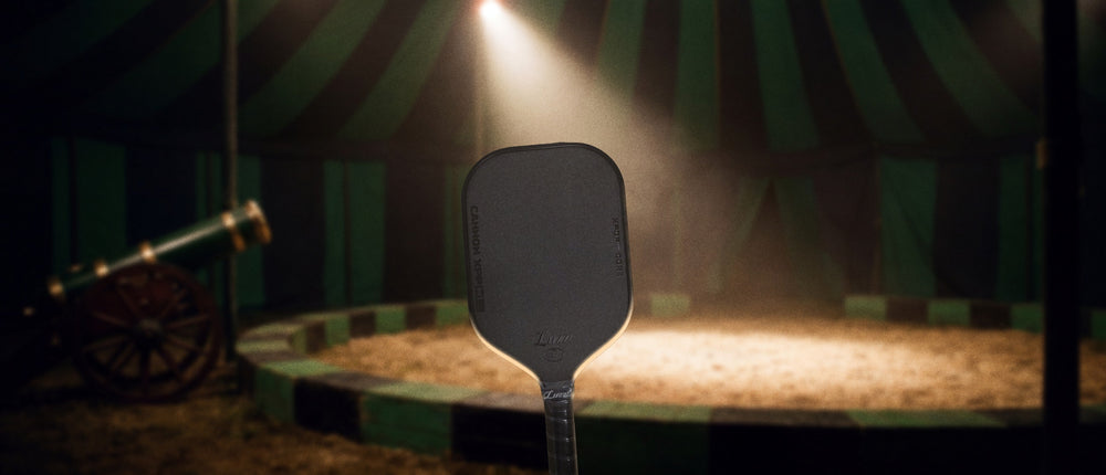 The Luzz Cannon XPP pickleball paddle standing in a spotlight in a green and black striped circus big top, an old circus cannon on the left