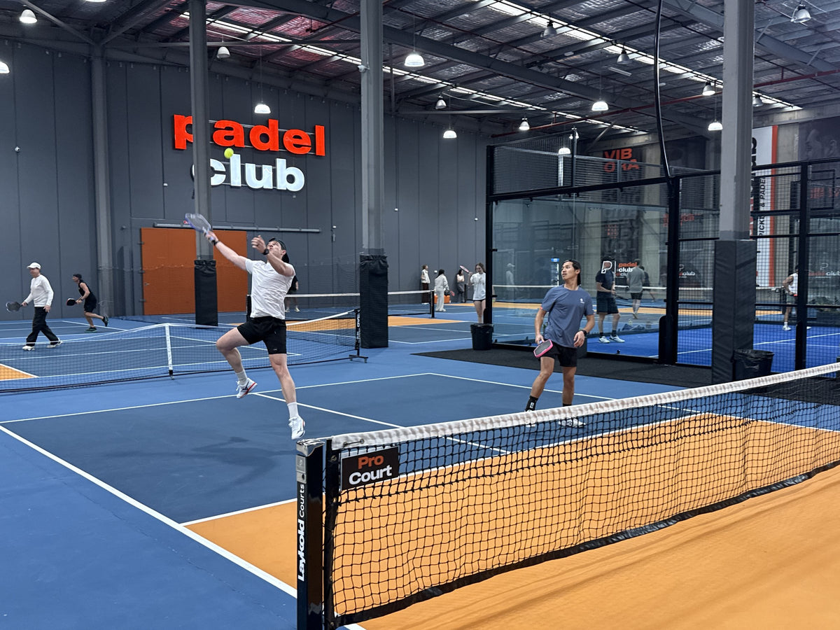 A player leaping for an overhead smash on an indoor pickleball court, with a social session playing on the courts behind.