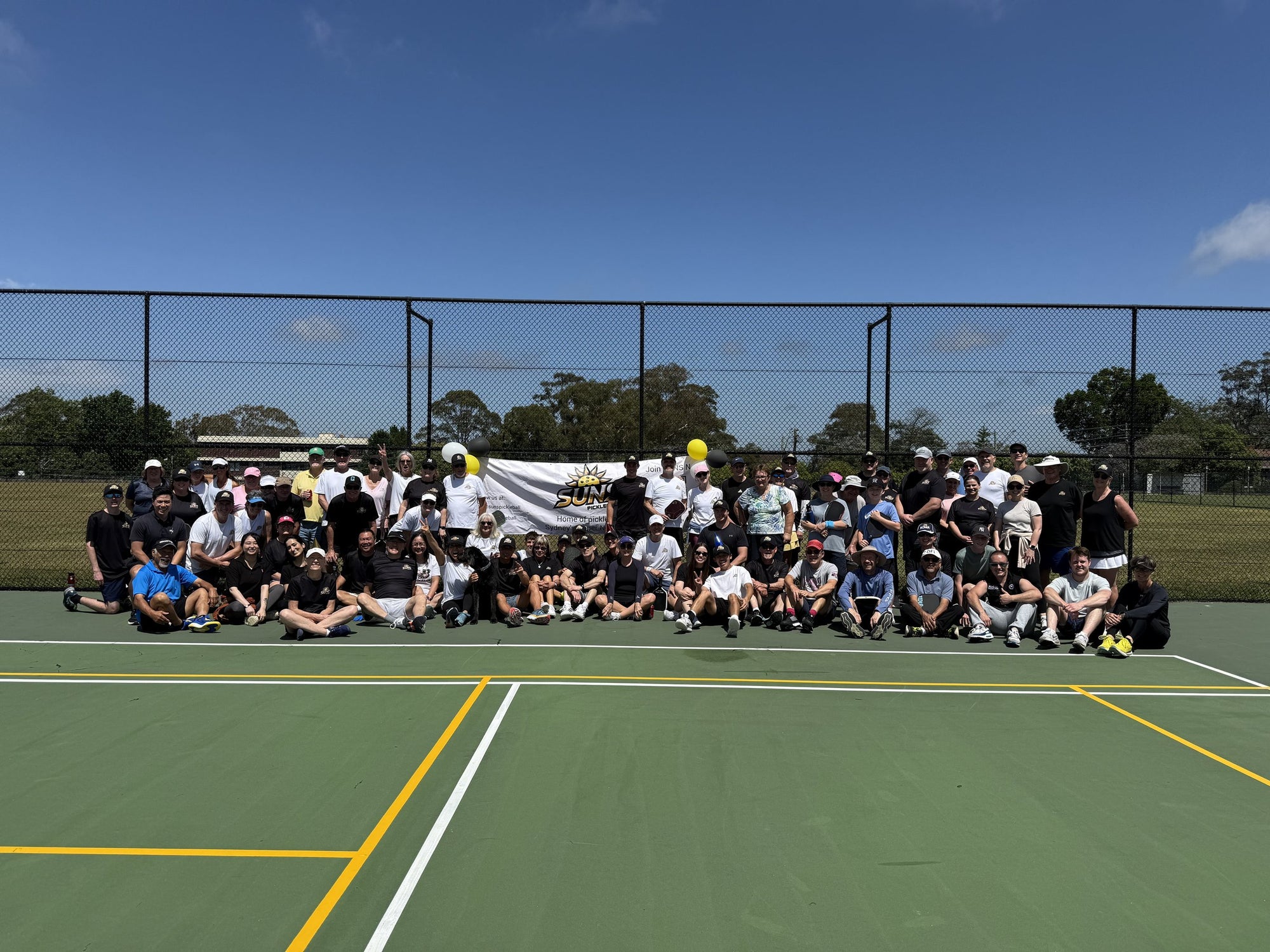 A pickleball club group photo on outdoor council courts, dozens of players sitting and standing in front of a club banner.