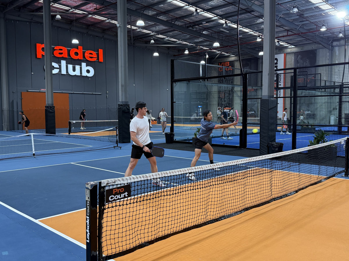 Two players rallying at the net on an indoor pickleball court at a commercial venue, with more courts in use behind them.